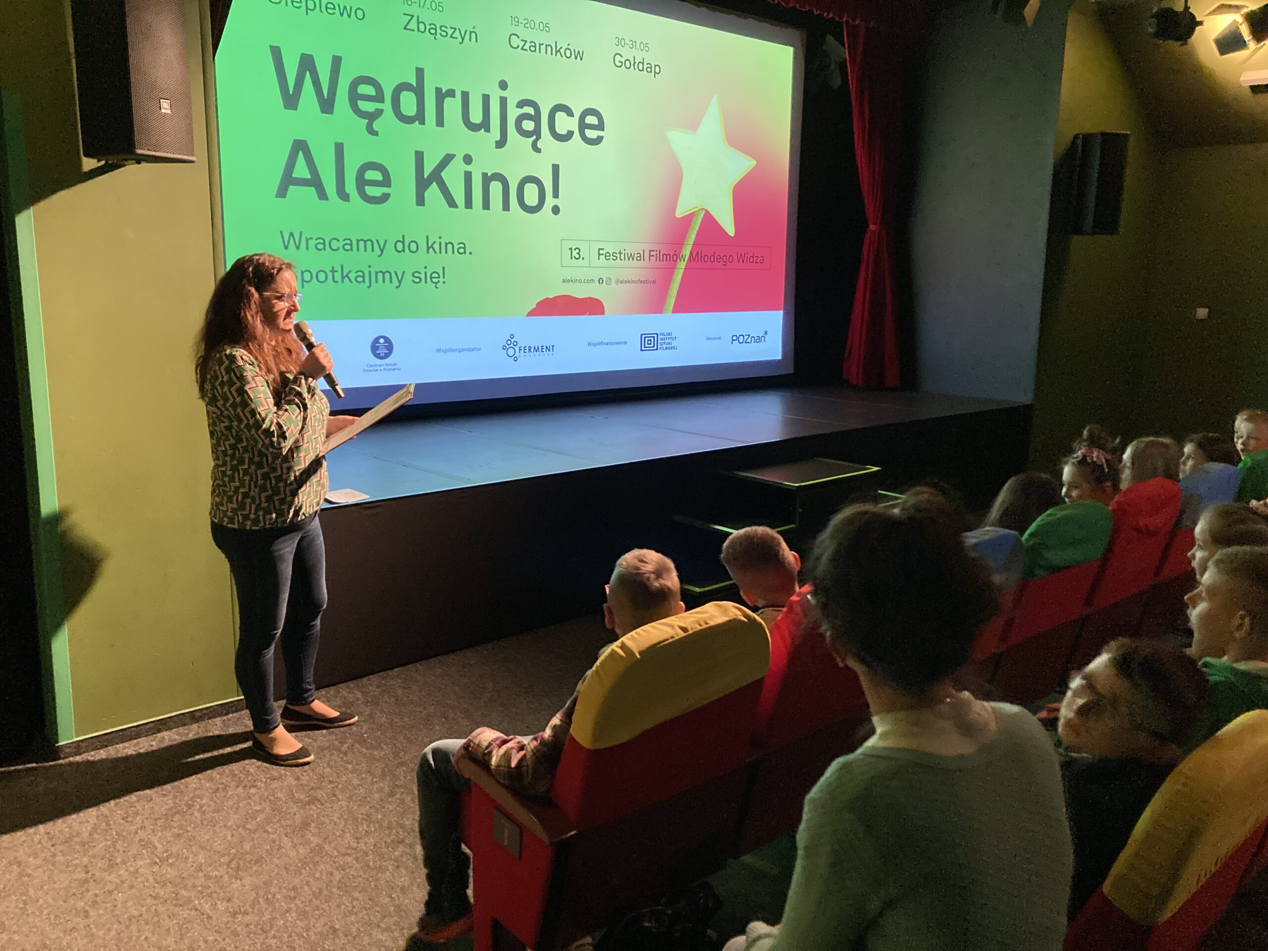 Agnieszka Michalczyk wita widzów w kinie na tle plakatu WAK na ekranie
