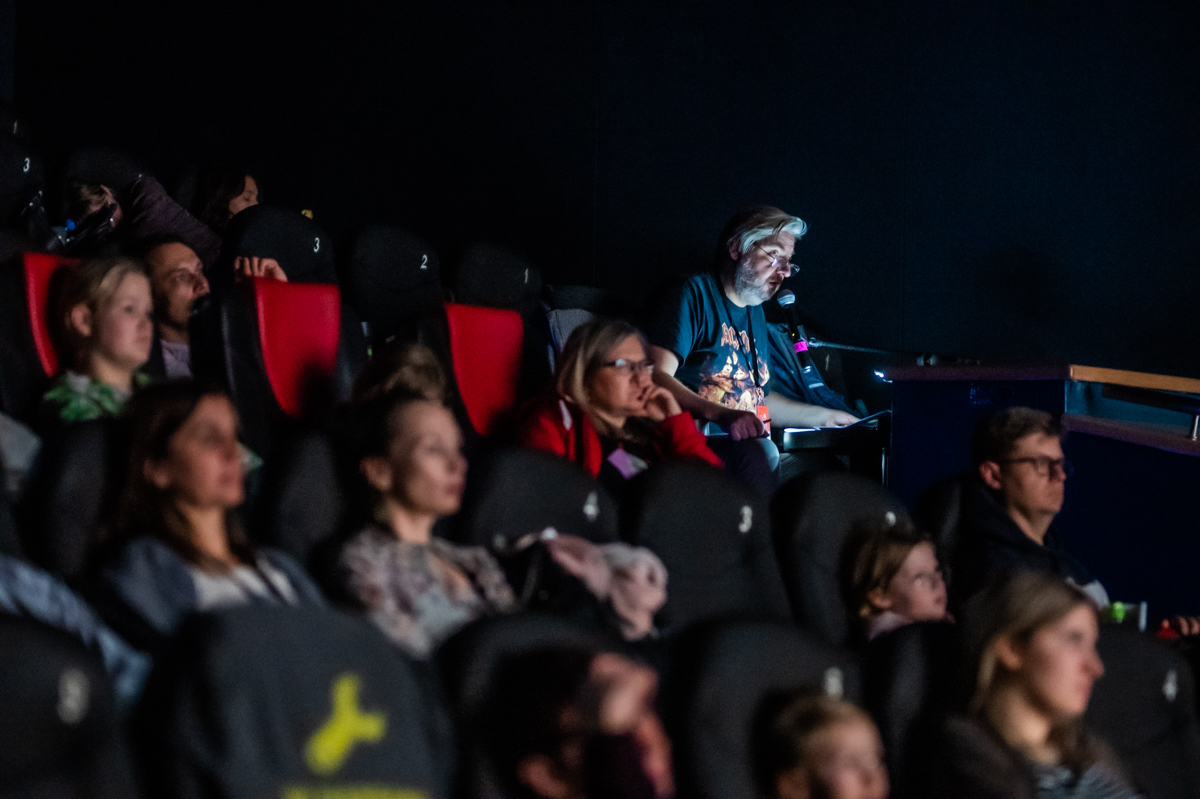 Publicznośc w ciemnej sali kinowej i lektor prowadzący audiodeskrypcję