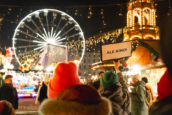 Osoba na ulicy w tłumie ludzi trzyma wysoko nad głową tabliczkę z napisem "Ale Kino!", w tle budki ozdobione światełkami i karuzela.