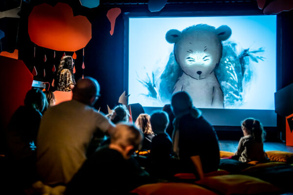 Grupa rodziców i dzieci siedzi na poduszkach i ogląda wyświetlany film animowany, na ekranie widać szarego misia.