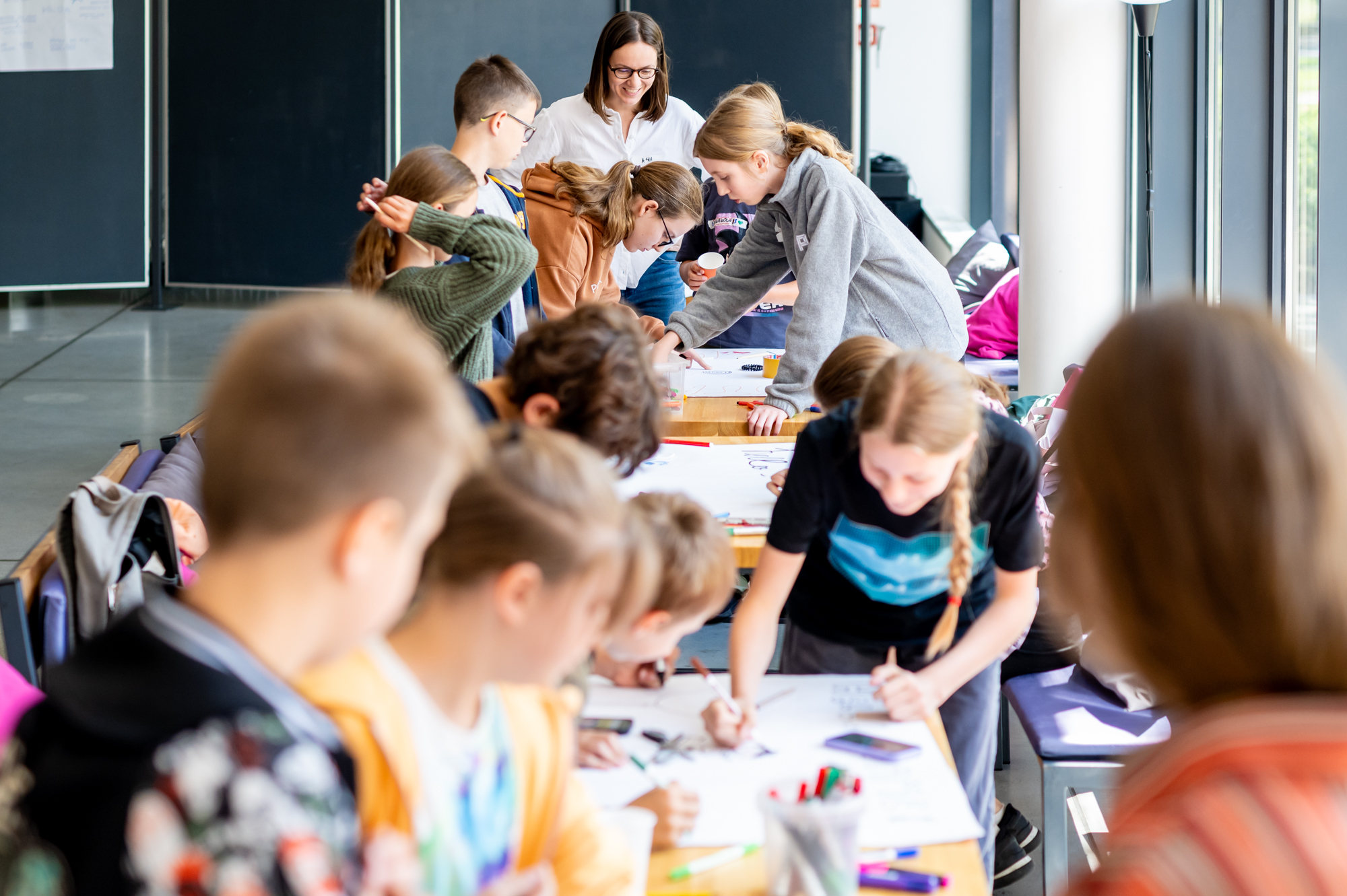 Grupa dzieci pracuje przy stołach, na których mają rozłożone kartki papieru, rysują i malują.