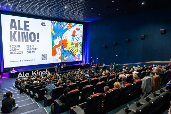 Sala kinowa widziana od tyłu, publiczność patrzy na ekran z plakatem festiwalu Ale Kino. Przed ekranem stoją cztery osoby, jedna mówi do mikrofonu.