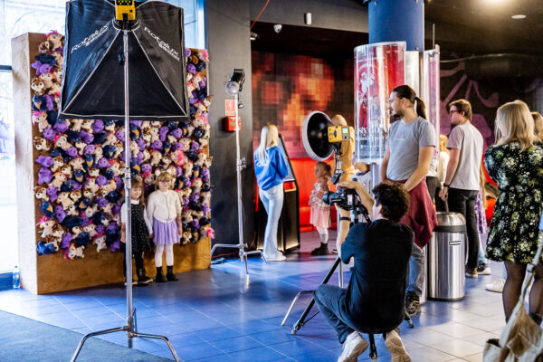 Na pierwszym planie widziany od tyłu fotograf trzymający aparat, w tle dwie dziewczynki pozujące na tle ścianki wypełnionej maskotkami.