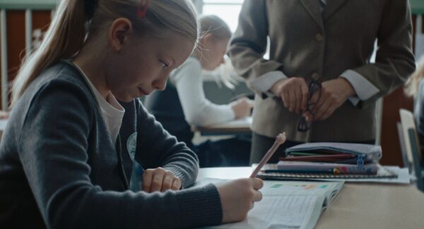 Dziewczynka w klasie siedzi przy biurku, ma przed sobą rozłożony zeszyt, jest spięta, obok niej stoi nauczycielka.