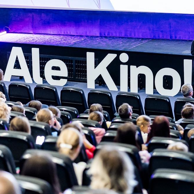 Sala kinowa, w fotelach siedzą ludzie. Przed nimi stoi napis z dużych, białych liter: Ale Kino!
