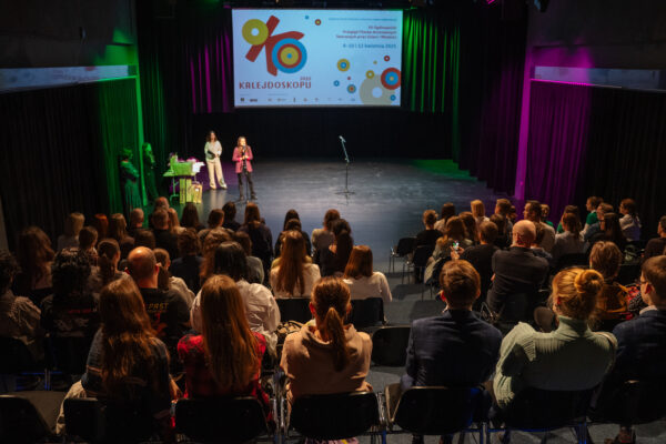 Sala, w której na widowni siedzą ludzie. Na scenie stoi kilka osób, jedna z nich mówi do mikrofonu. W tle duży, jasny ekran z kolorowym logo projektu Oko Kalejdoskopu.