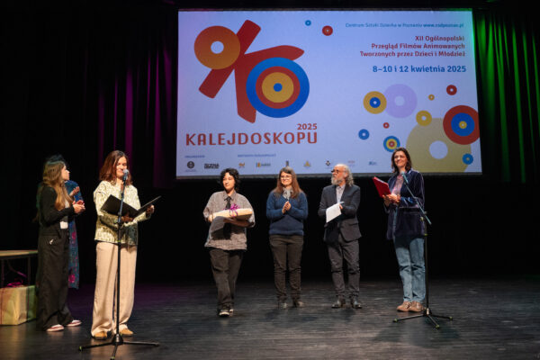 Kilka osób stoi w rzędzie obok siebie, przed nimi dwa mikrofony. Część osób bije brawo. Za nimi jasny ekran z kolorowym logo projektu Oko Kalejdoskopu.