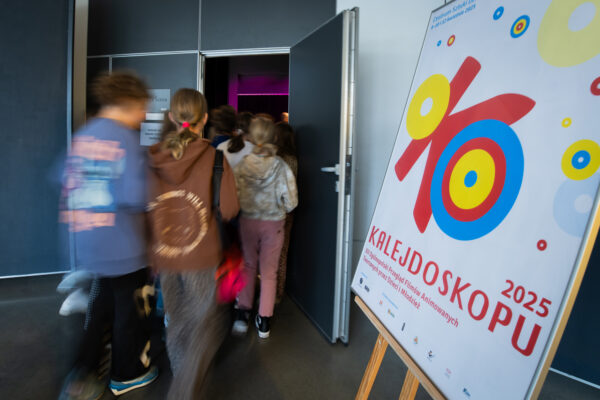 Grupa dzieci wchodzi do ciemnej sali. Postaci są rozmazane. Przy drzwiach ustawiony plakat z logo projektu Oko Kalejdoskopu 2025.