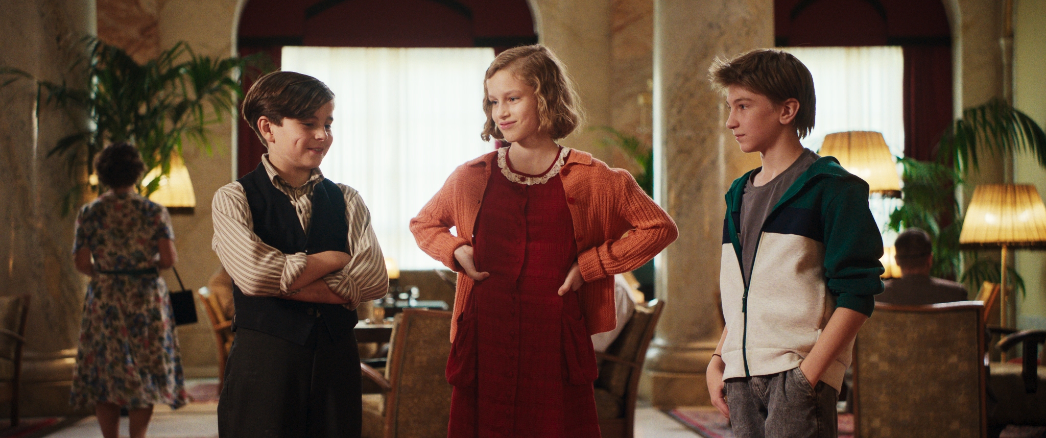Kadr z filmu. Dwóch chłopców i dziewczynka stoją razem, dwoje z nich się uśmiech i ma na sobie ubrania z lat trzydziestych dwudziestego wieku. Stoją w eleganckim holu z marmurowymi kolumnami.