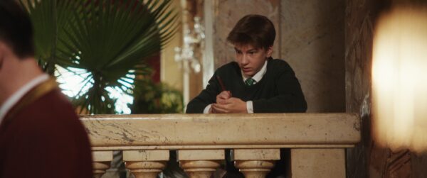 Kadr z filmu. Młody chłopiec opiera się o balustradę i zapisuje coś ołówkiem na swojej dłoni.