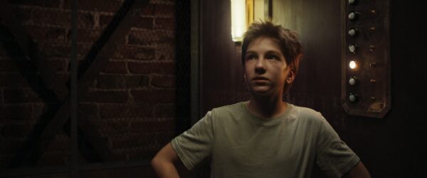 Kadr z filmu. Młody chłopiec stoi w starodawnej widzie. Obok niego ściana z dużymi przyciskami, jeden z nich świeci się.