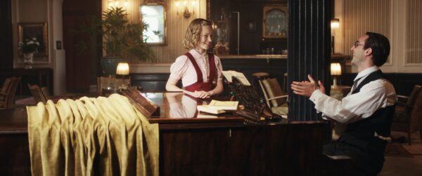 Kadr z filmu. Młoda, uśmiechnięta dziewczynka stoi przy pianinie. Patrz na mężczyznę, który siedzi przy pianinie, śmieje się i klaszcze w dłonie. Noszą stroje z lat trzydziestych dwudziestego wieku.