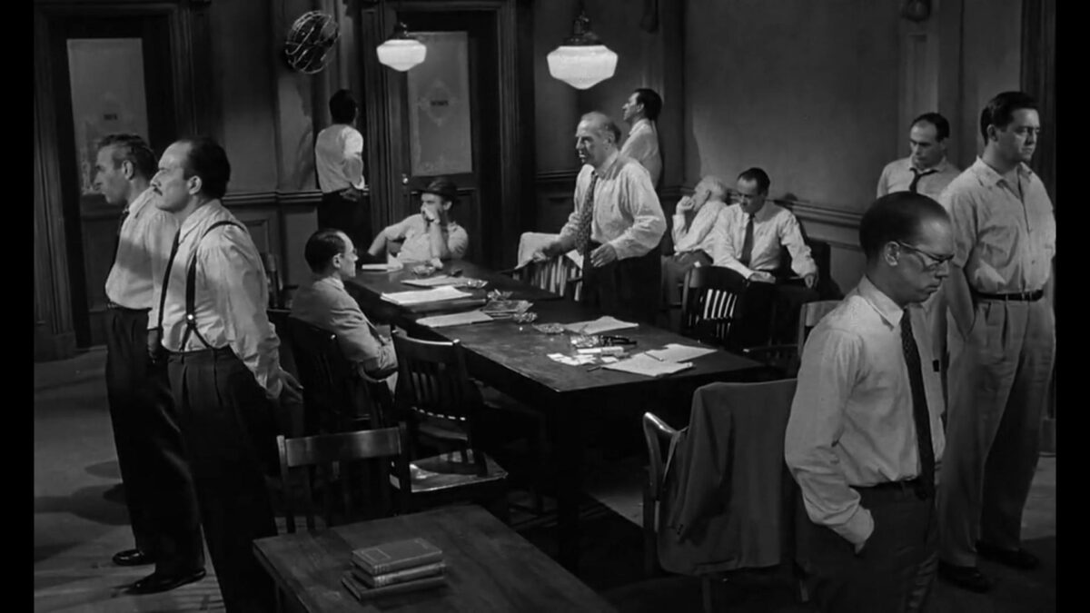 Osoby w garniturach siedzą przy stole, niektórzy patrzą przez okno obok