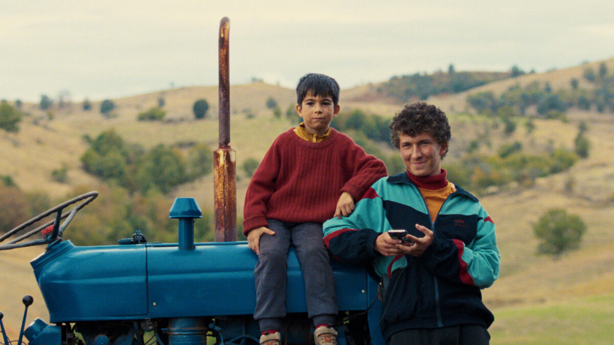 chłopczyk w czerwonym swetrze siedzi na traktorze, obok niego stoi uśmiechnięty mężczyzna w kurtce