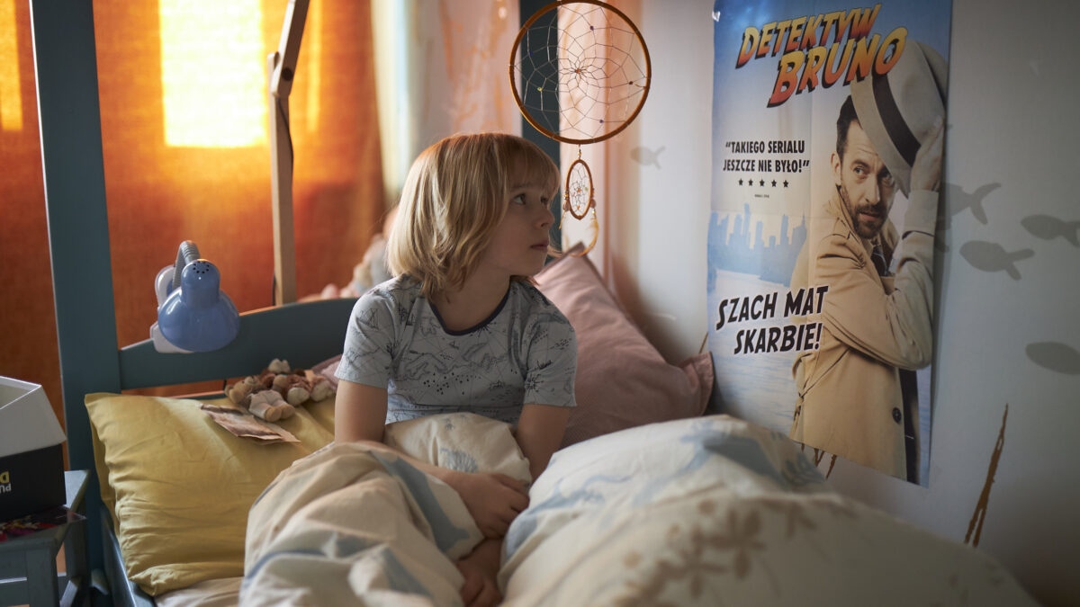 Chłopiec siedzi na łóżku w pokoju ozdobionym plakatem filmowym, w pomarańczowym świetle poranka