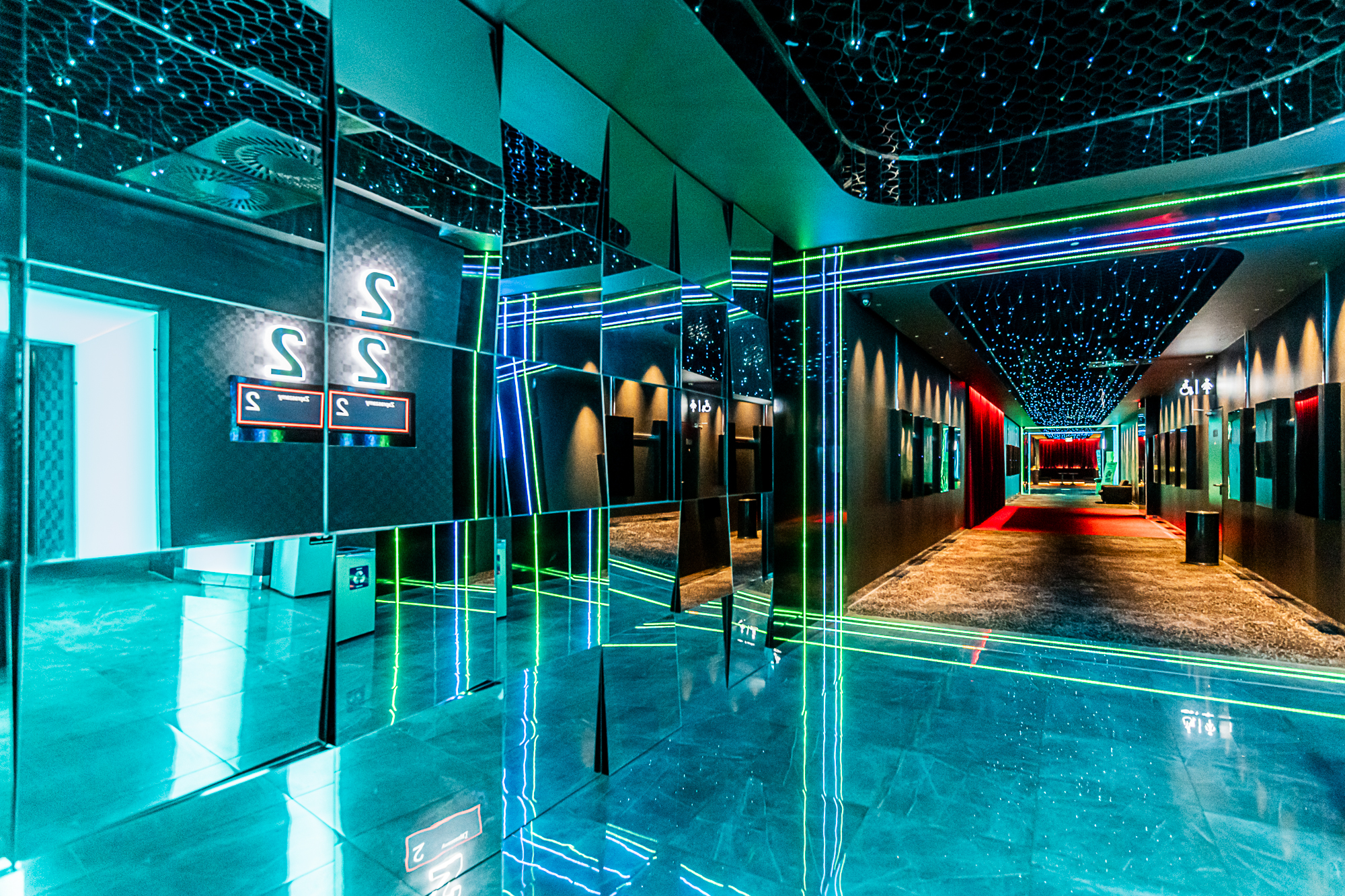 Korytarz z jedną ze ścian wyłożoną lustrzanymi kaflami. Część podłogi jest wyłożona kafelkami, część wykładziną. Na suficie małe, błyszczące światełka.