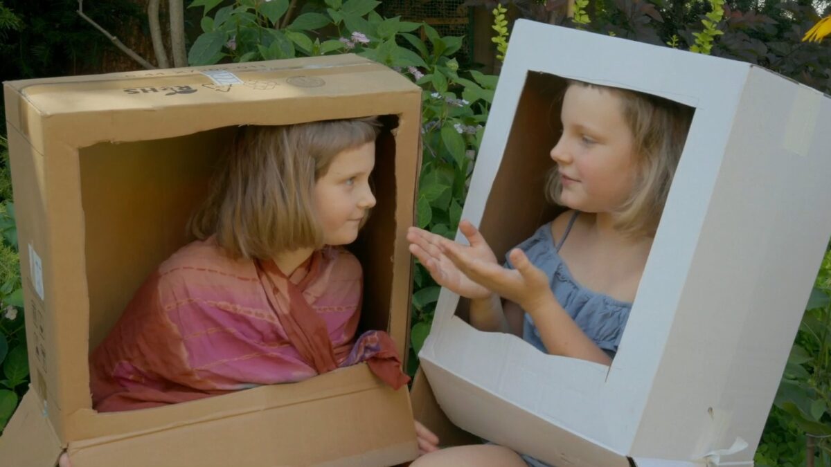 dwie dziewczynki siedzą w kartonowych pudełkach przypominających telewizory i rozmawiają ze sobą