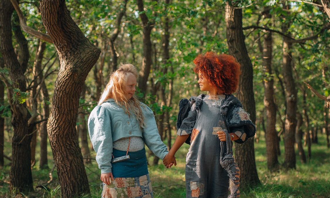 dwie dziewczynki o rudych i jasnych włosach trzymają się za ręce w lesie