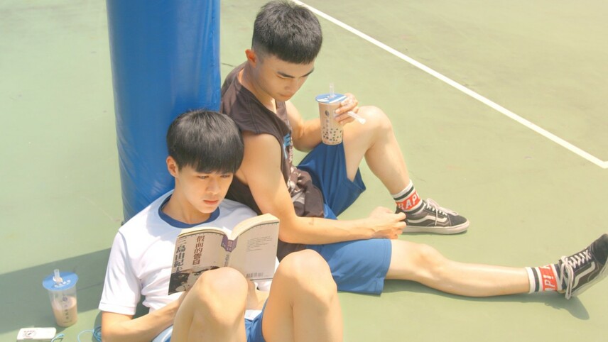 Dwaj chłopcy siedzą przy słupie na terenie sportowym — jeden czyta książkę, a drugi trzyma napój i zagląda do książki kolegi