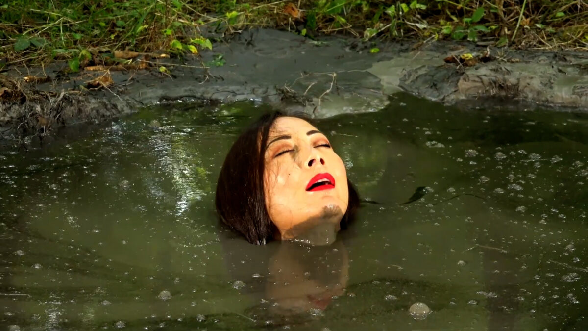 Kobieta o ciemnych włosach i czerwonych ustach leży w błocie z zamkniętymi oczami