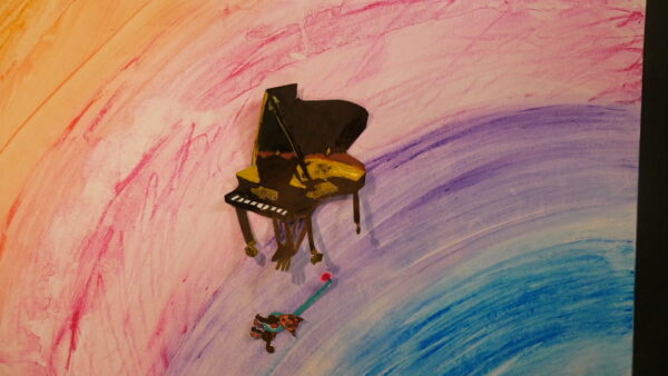 Kadr z filmu animowanego, papierowa makieta fortepianu stoi na tle kolorowego, malowanego tła z przejściami różu i błękitu, obok leży mała postać.