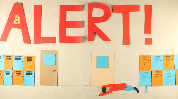Kadr z filmu animowanego, duży czerwony napis „ALERT!” znajduje się nad rzędem kolorowych drzwi i szafek, a na podłodze leży postać wyciągająca ręce do przodu.