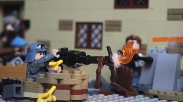 Kadr z filmu animowanego, figurka żołnierza z klocków stoi za murkiem i obsługuje karabin maszynowy, z którego wydobywa się ogień.