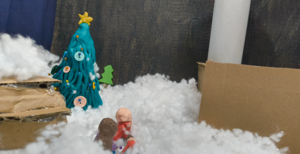 Kadr z filmu animowanego, plastelinowa choinka ozdobiona guzikami i gwiazdką stoi wśród białej waty imitującej śnieg, obok znajdują się kartonowe elementy.