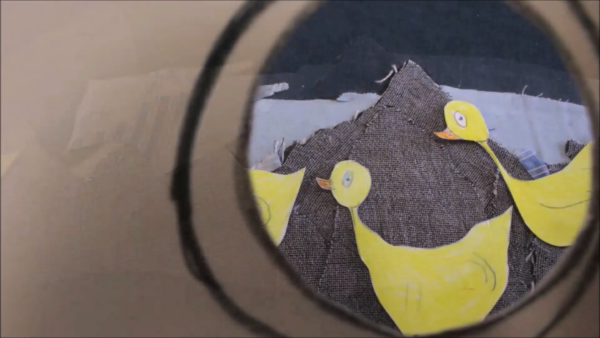 Kadr z filmu animowanego, dwie papierowe postaci kaczki stoją na tle brązowej tkaniny, uchwycone są w kadrze z okrągłego wizjera.