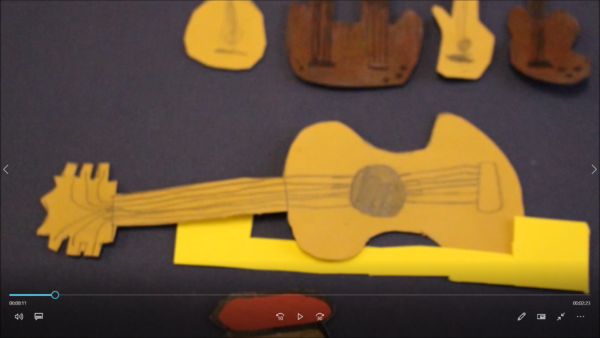 Kadr z filmu animowanego, papierowe instrumenty muzyczne, w tym gitara i inne elementy instrumentów, ułożone są na ciemnym tle.