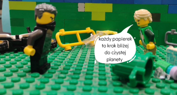 Kadr z filmu animowanego, dwie figurki z klocków stoją na zielonej powierzchni obok rozrzuconych elementów i przewróconego kosza, jedna z postaci mówi w dymku tekst o sprzątaniu planety.