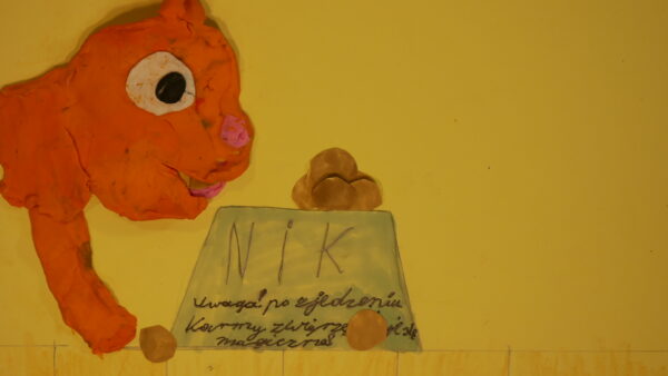 Kadr z filmu animowanego, plastelinowa pomarańczowa postać pochyla się nad miską z napisem, obok leżą kulki jedzenia.