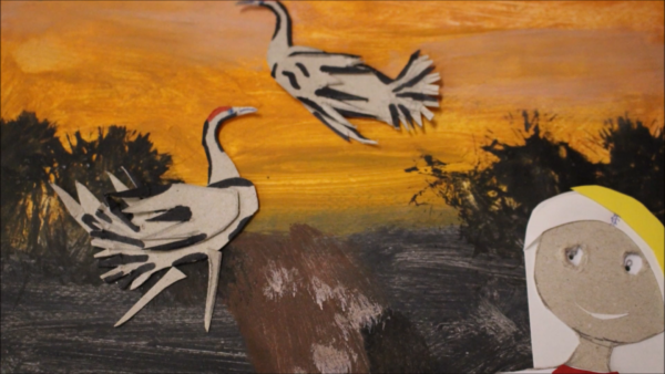 Kadr z filmu animowanego, dwie białe papierowe ptaki lecą nad ciemnym krajobrazem przy pomarańczowym zachodzie słońca, obok stoi postać w jasnym nakryciu głowy