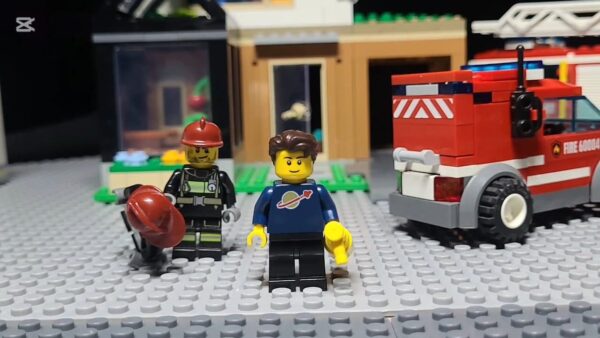 Kadr z filmu animowanego, figurki z klocków przedstawiają strażaka i chłopca stojących przed budynkiem i czerwonym wozem strażackim.