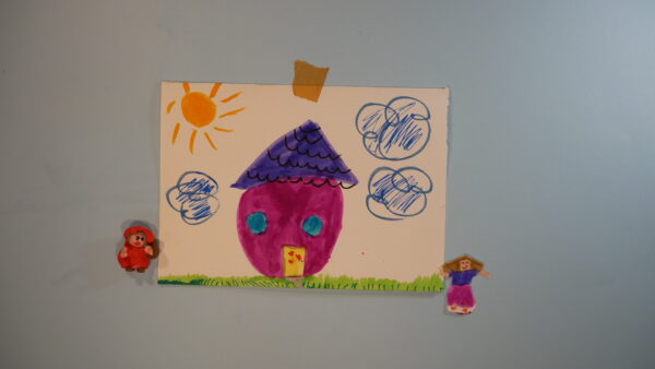 Kadr z filmu animowanego przedstawia dziecięcy rysunek domu z kolorowymi elementami, przyczepiony do ściany.