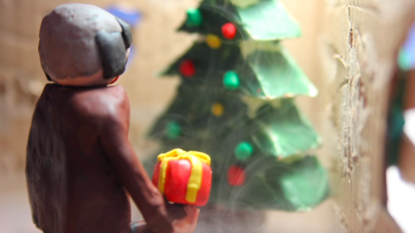 Kadr z filmu animowanego, postać z plasteliny trzyma w ręku prezent i stoi przed ozdobioną choinką w pomieszczeniu.