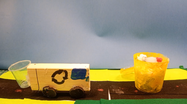 Kadr z filmu animowanego ukazuje ręcznie wykonany model pojazdu stojącego obok małego pojemnika na kolorowym podłożu.