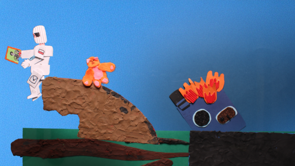 Kadr z filmu animowanego, papierowa postać siedzi na brzegu ziemi z pluszowym misiem, a obok widoczna jest płonąca konstrukcja.