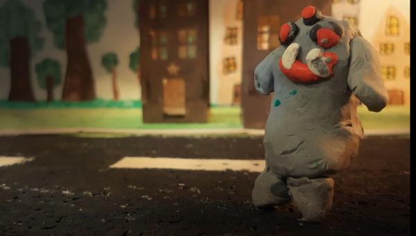 Kadr z filmu animowanego, plastelinowa postać o nieregularnym kształcie stoi na ulicy, a w tle widać budynki i drzewa.