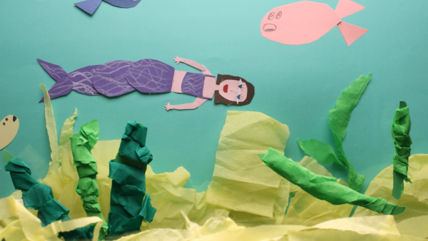 Kadr z filmu animowanego, papierowa postać syreny unosi się w wodzie wśród zielonych roślin i ryb.