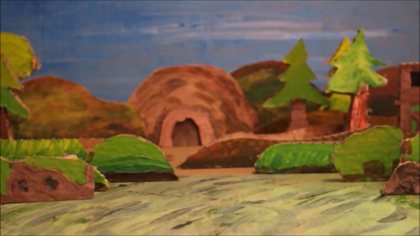 Kadr z filmu animowanego, papierowy krajobraz z drzewami i wzgórzami prowadzi do wejścia do jaskini znajdującej się pośrodku sceny.