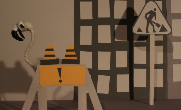 Kadr z filmu animowanego, papierowa makieta miasta przedstawia znak robót drogowych, dwa pachołki oraz sylwetkę ptaka stojącego obok.
