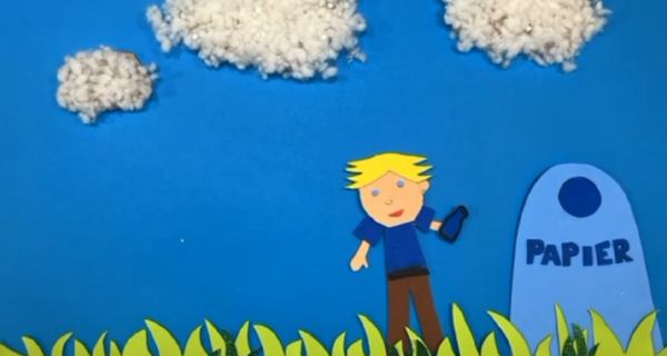 Kadr z filmu animowanego, wycięta z papieru postać chłopca stoi na trawie i wrzuca przedmiot do pojemnika z napisem „papier”, na tle nieba z chmurami.