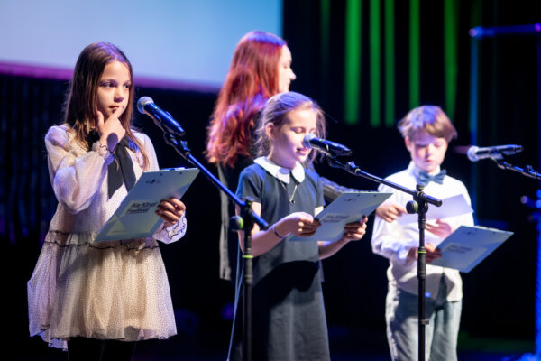 dwie dziewczynki i chłopiec stoją na scenie i mówią do mikrofonu