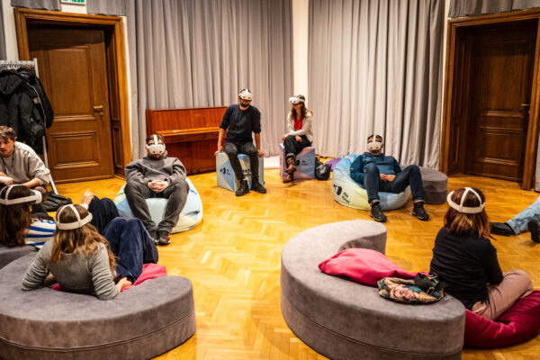 Grupa dzieci i dorosłych siedzi na kolorowych pufach i kostkach z napisem: Ale Kino! Na głowach mają okulary VR.