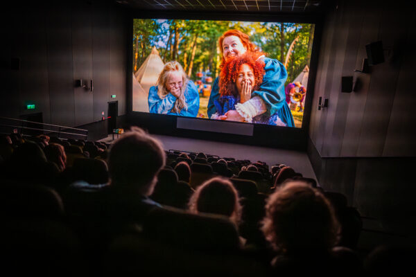 Sala kinowa wypełniona publicznością. Na ekranie kadr z filmu: kobieta w rudych włosach obejmuje rudowłosą dziewczynkę. Obok druga dziewczynka w długich jasnych włosach, wszystkie trzy chichoczą.