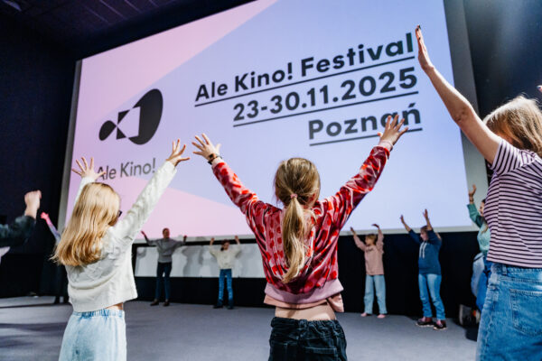 Dzieci w kole stoją z wysoko uniesionymi rękoma. Za nimi duży ekran kinowy z planszą z napisami: Ale Kino! Festival 23-30.11.2025 Poznań